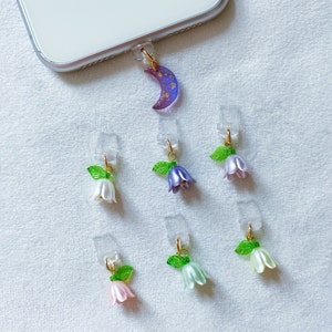 Puede incluir: Un conjunto de colgantes para teléfono con forma de luna creciente y flores en forma de campana en varios colores, incluyendo blanco, morado, rosa y verde. Los colgantes están unidos a soportes de plástico transparente.