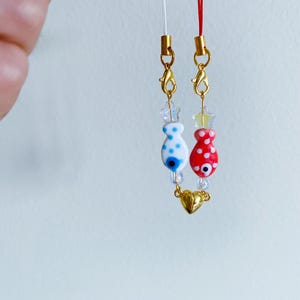 May include: Two gold charm necklaces with white and red fish charms with blue evil eye details. The charms are connected by a gold heart charm.