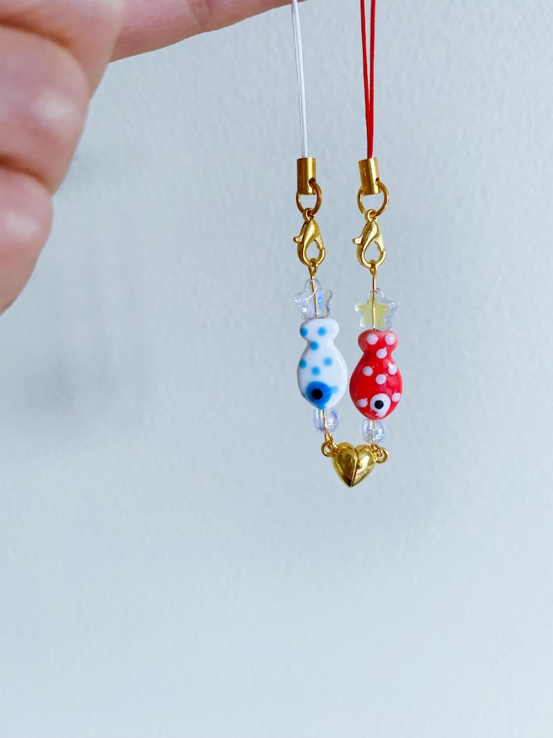 May include: Two gold charm necklaces with white and red fish charms with blue evil eye details. The charms are connected by a gold heart charm.