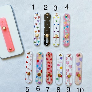 May include: Ten different phone grips with heart designs. The grips are made of clear plastic with a variety of colorful heart patterns. The grips have a gold ring on the back for easy holding.