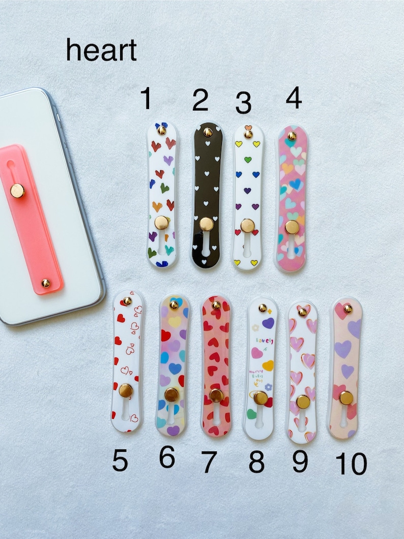 May include: Ten different phone grips with heart designs. The grips are made of clear plastic with a variety of colorful heart patterns. The grips have a gold ring on the back for easy holding.
