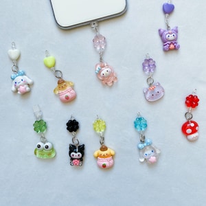 Puede incluir: Una colección de 10 abalorios de teléfono coloridos con varios personajes de dibujos animados, incluyendo una rana, un hongo, un gato, un conejito y un perro. Los abalorios están unidos a cuentas transparentes y cadenas plateadas.