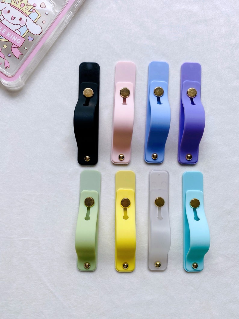 May include: Eight phone grips in various colors, including black, pink, light blue, purple, light green, yellow, white, and light blue. The grips are made of silicone and have a gold metal button.