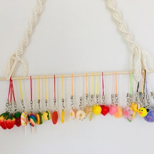 May include: A collection of colorful, miniature food-themed charms hanging from a wooden dowel. The charms include a strawberry, lollipop, hamburger, orange slice, egg, donut, lemon, heart, orange, flower, duck, and a fuzzy purple ball.