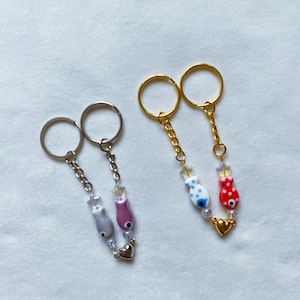 May include: Two sets of keychains with silver and gold rings. Each set has two charms: a small heart and a fish with an evil eye. One set has a pink and gray fish, the other has a red and white fish.