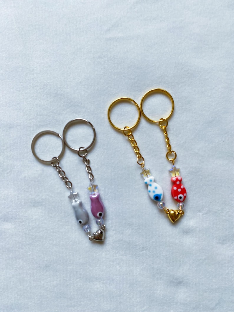 May include: Two sets of keychains with silver and gold rings. Each set has two charms: a small heart and a fish with an evil eye. One set has a pink and gray fish, the other has a red and white fish.