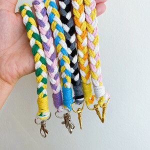 May include: Six colorful braided keychains with gold metal clasps. Each keychain is made of two colors of yarn, braided together in a diagonal pattern. The colors include green, yellow, pink, purple, blue, and gray.