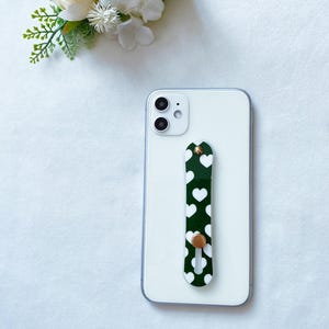 May include: A white smartphone with a green phone grip with white hearts. The grip has a gold button.