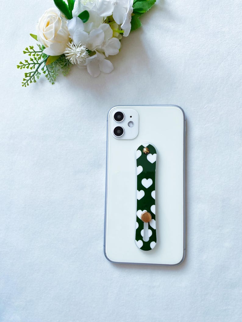May include: A white smartphone with a green phone grip with white hearts. The grip has a gold button.