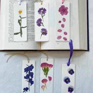 May include: Six rectangular bookmarks featuring pressed flowers. Three bookmarks are placed on an open book, and three are below it. They display various floral designs including a white daisy, purple asters, pink petals, blue delphiniums, pink carnations, and purple pansies, each with a ribbon or twine tie.
