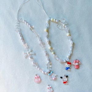 May include: Two white beaded phone charms with various charms including butterflies, a bow, and cartoon characters. The charms are attached to white strings.