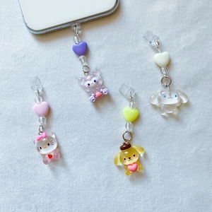 May include: Four phone charms featuring cartoon characters. The charms are made of clear plastic with colourful accents. The characters include a pink Hello Kitty, a yellow dog with a brown hat, a purple cat with a pink bow, and a white dog with blue eyes.