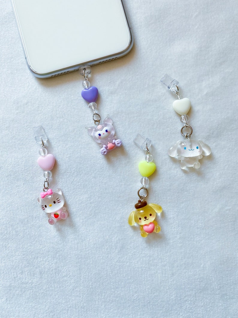 May include: Four phone charms featuring cartoon characters. The charms are made of clear plastic with colourful accents. The characters include a pink Hello Kitty, a yellow dog with a brown hat, a purple cat with a pink bow, and a white dog with blue eyes.