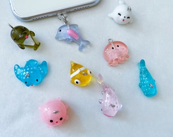 May include: A set of nine colorful sea creature phone charms. The charms include a dolphin, an octopus, a seal, a stingray, a clownfish, a whale shark, a manta ray, and two pink sea creatures.