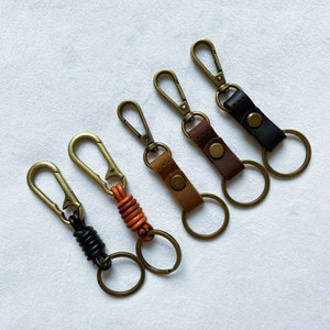 May include: Five keychains with different styles. Each keychain has a metal clasp and a ring. One keychain has a black leather strap, one has a brown leather strap, one has a tan leather strap, and two have a braided leather strap.