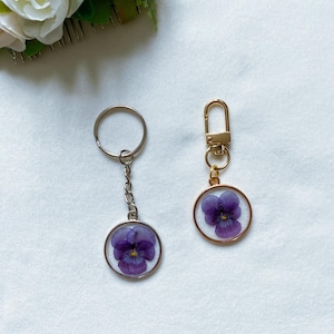 May include: Two keychains with a purple flower pressed inside a clear resin circle. One keychain has a silver ring and chain, the other has a gold ring and chain.