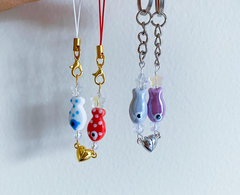 May include: Two colorful fish charm keychains with gold and silver accents. One keychain features a white fish with blue spots and a red fish with white spots. The other keychain features a light purple fish with a silver heart charm.