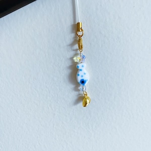 May include: A white and blue charm with a blue evil eye design and white polka dots hangs from a gold chain with a gold bell charm. The charm is attached to a white string.