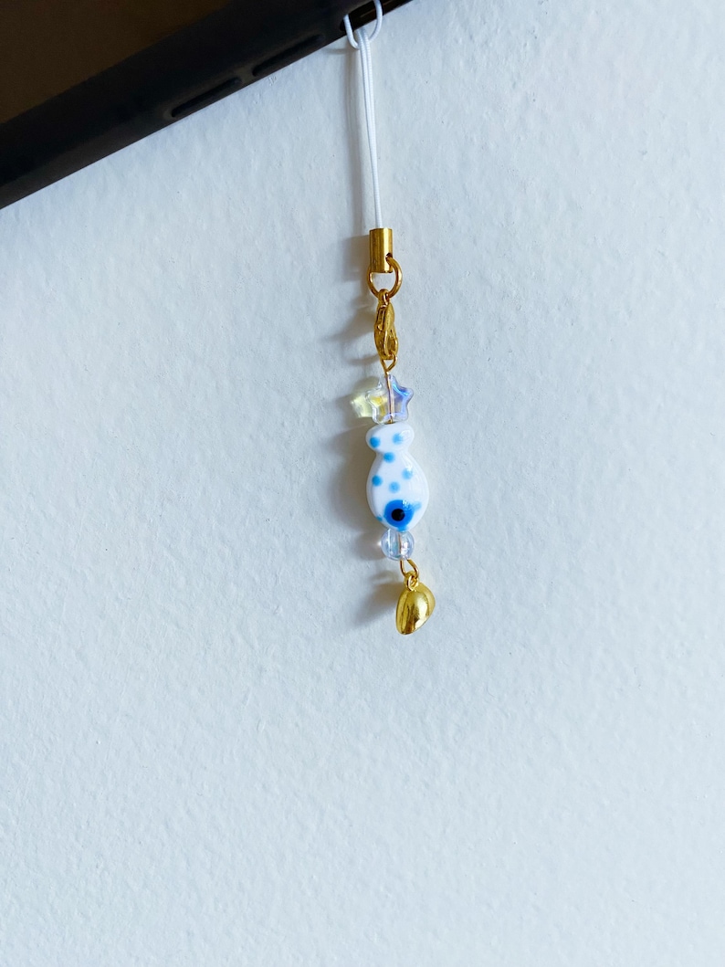 May include: A white and blue charm with a blue evil eye design and white polka dots hangs from a gold chain with a gold bell charm. The charm is attached to a white string.