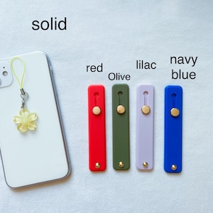 May include: A white smartphone with a yellow flower charm attached to it.  The phone is next to five phone grips in different colors: red, olive green, lilac, navy blue, and solid.