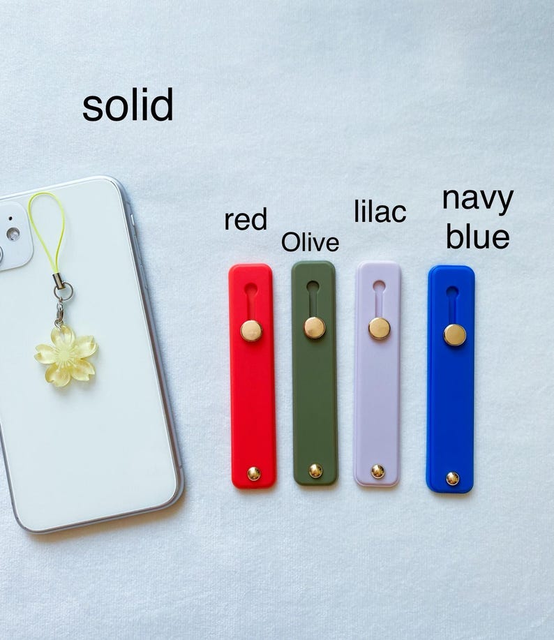 May include: A white smartphone with a yellow flower charm attached to it.  The phone is next to five phone grips in different colors: red, olive green, lilac, navy blue, and solid.