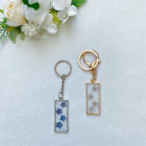 Könnte beinhalten: Zwei Schlüsselanhänger mit rechteckigen Rahmen, die gepresste Blumen enthalten. Ein Schlüsselanhänger hat blaue Blumen und einen silbernen Rahmen, der andere hat weiße Blumen und einen goldenen Rahmen.