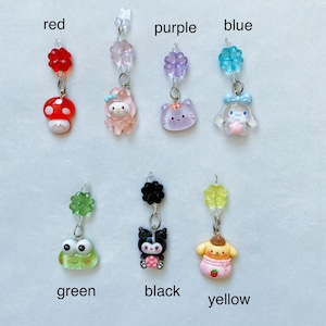 May include: A set of 10 charm pendants featuring various colours and characters. The charms are attached to a clear string and include a red mushroom, a pink Hello Kitty, a purple cat, a blue clover, a green frog, a black Kuromi, a yellow Pompompurin, a pink flower, a purple flower, and a blue flower.