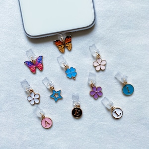Op de afbeelding: Assortiment van telefooncharms met een vlinder, een bloem, een klavertjevier, een ster, een letter en een cirkel met een letter. De charms zijn gemaakt van metaal en hebben een gouden afwerking.