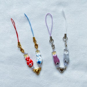 May include: Three phone charms with different colored strings and fish charms. The charms have a gold heart charm and clear beads. The strings are red, blue, and purple.