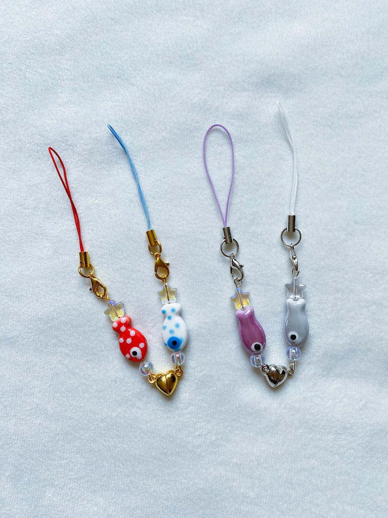 May include: Three phone charms with different colored strings and fish charms. The charms have a gold heart charm and clear beads. The strings are red, blue, and purple.