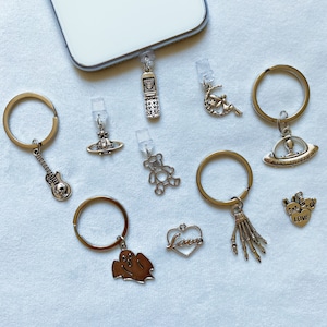 May include: A set of 10 silver charm phone charms. The charms include a ghost, a skeleton hand, a heart with the word "love" on it, a teddy bear, a guitar, a UFO, a crescent moon with a fairy, a phone, and a orb with a star.
