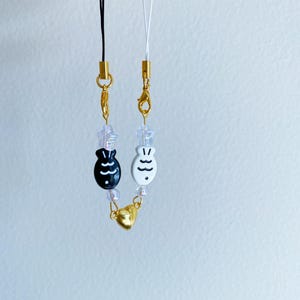 May include: Two phone straps with gold charms and black and white fish charms. The straps are made of clear beads and gold chains.