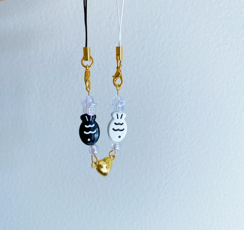 May include: Two phone straps with gold charms and black and white fish charms. The straps are made of clear beads and gold chains.