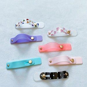 May include: A set of seven phone grips with different designs. The grips are made of clear plastic with various patterns, including hearts, butterflies, and polka dots. Each grip has a gold button.