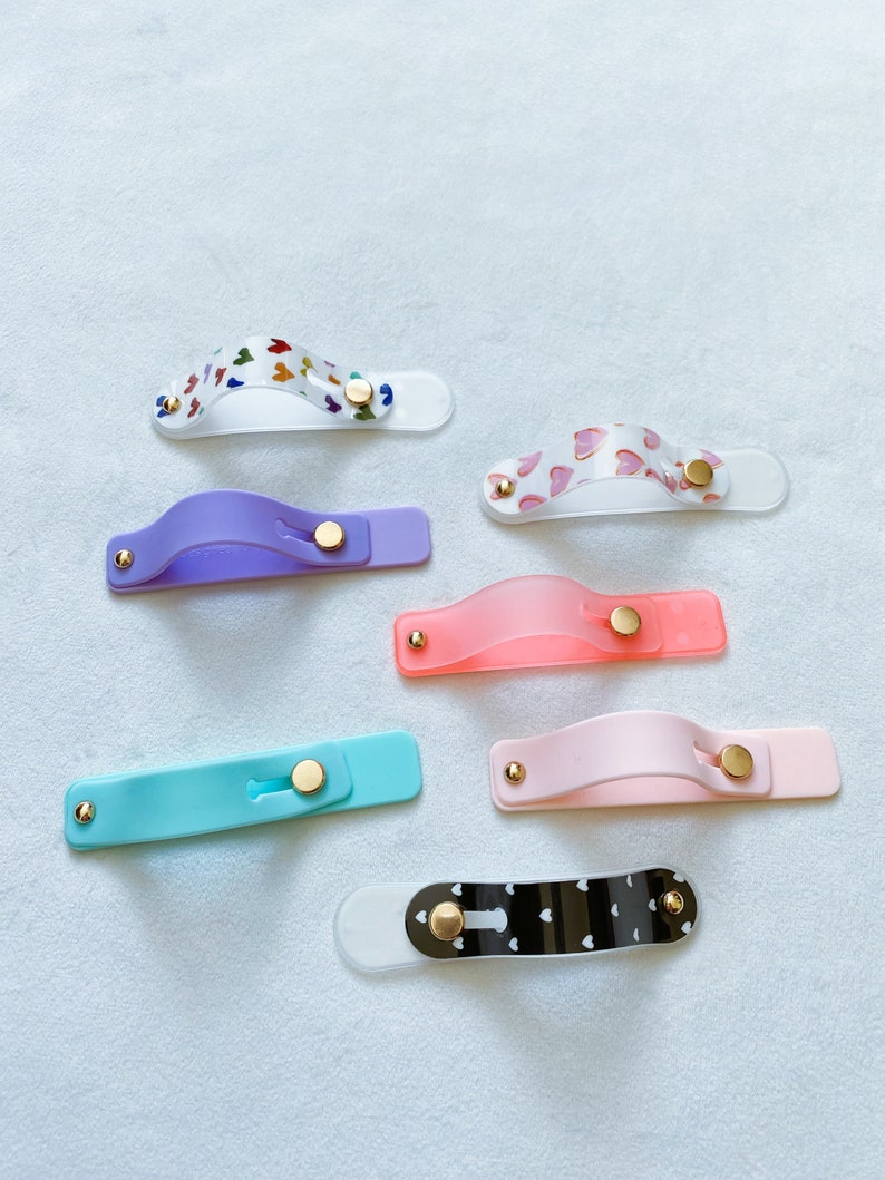 May include: A set of seven phone grips with different designs. The grips are made of clear plastic with various patterns, including hearts, butterflies, and polka dots. Each grip has a gold button.