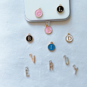May include: A set of alphabet letter charms in various colours and styles. The charms are made of metal and have a gold finish. The letters are arranged in a random order on a white surface.