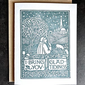 May include: A teal and white Christmas card with a pastoral scene. The card features a shepherd with a staff, sheep, a tree, and a building. The text reads "I Bring You Glad Tidings."
