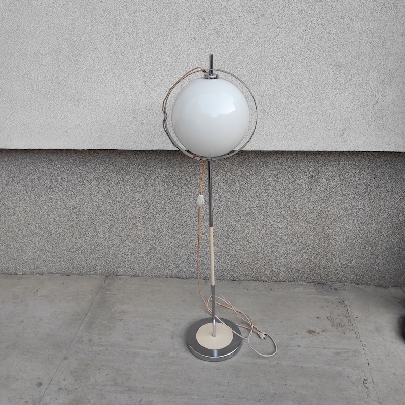 May include: A white globe lamp with a chrome stand and a white cord. The lamp is on a gray concrete floor.