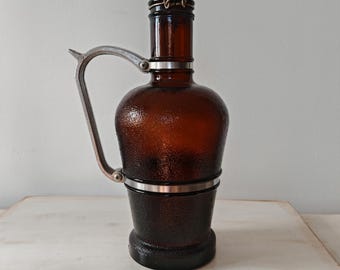 May include: A brown glass bottle with a textured surface, a metal handle, and a hinged metal lid. The bottle has a bulbous body and a cylindrical base. The handle is attached to the bottle with metal rings.