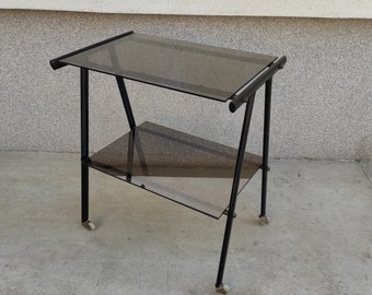 May include: A black metal two-tiered serving cart with glass shelves. The cart has a dark brown tint and is on wheels.