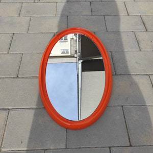 Op de afbeelding: Een ovale spiegel met een feloranje frame. De spiegel reflecteert een gebouw en de lucht.