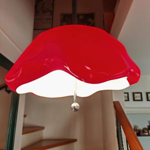 May include: A vibrant red pendant light with a unique, organic shape. The light fixture has a glossy finish and a white interior, suspended from a black cord. A small, silver sphere hangs below the light. The light is above a wooden staircase.