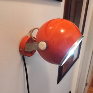 Puede incluir: Una lámpara de pared de color naranja brillante con una cabeza redonda giratoria y un brazo de metal plateado. La lámpara está cableada y tiene una perilla blanca en el brazo.