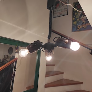 May include: A black metal ceiling light fixture with three light bulbs. The light fixture has a long, flexible cord and is hanging from the ceiling. The light bulbs are all turned on and are illuminating the room.