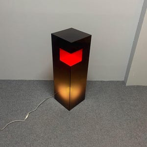 May include: A modern black floor lamp with a red chevron design. The lamp has a rectangular shape and emits a warm glow. A white power cord extends from the base.
