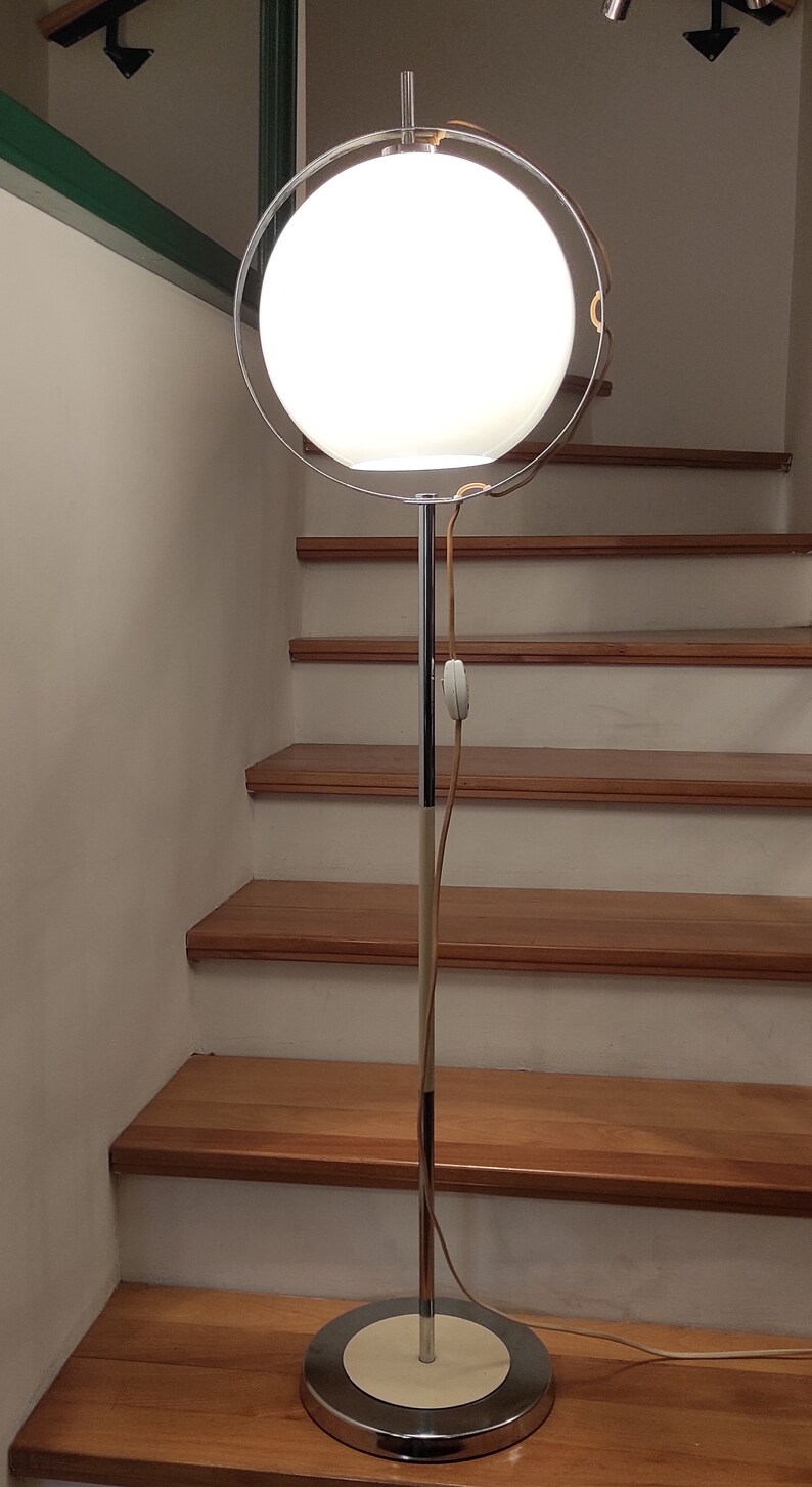 May include: A chrome floor lamp with a white globe shade and a circular chrome frame. The lamp is on a wooden floor with stairs in the background.