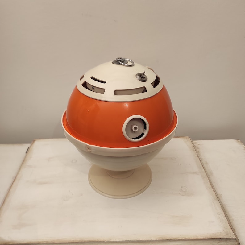 May include: A vintage orange and white plastic table lamp with a round base and a globe-shaped shade. The shade has a series of holes and a small metal ring on top.