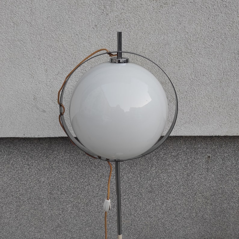 May include: A white globe lamp with a chrome frame and a brown cord. The lamp is on a white wall.