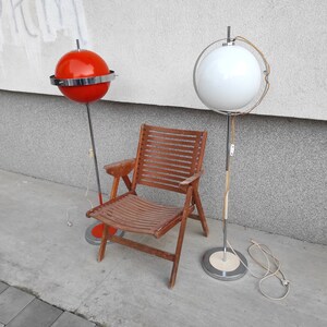 May include: Two mid-century modern floor lamps with white and red globe shades. The lamps are next to a wooden chair with a slatted back.