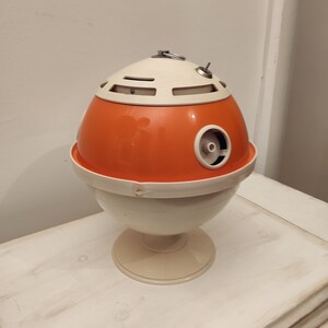 May include: Orange and white egg-shaped vintage ashtray with a metal ring on top. The ashtray has a round base and a small hole on the side.
